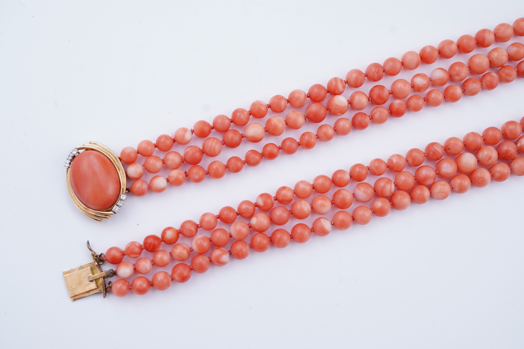 A coral necklace, composed of three strands of coral beads measuring approximately 5.1-6.6mm diameter, to a gold clasp set with a coral cabochon and brilliant-cut diamonds, mounted in gold, length 45.5cm, stamped 14K, ca